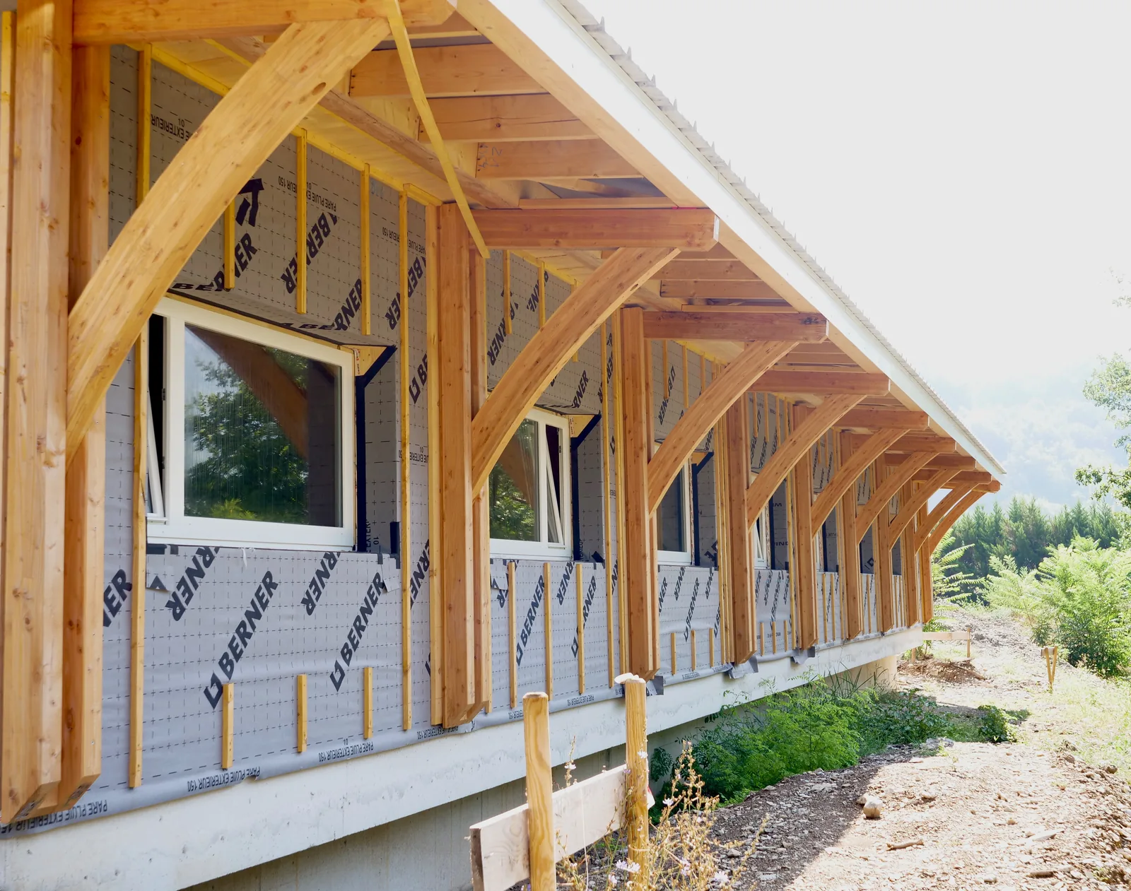 La charpente en bois de l’atelier en construction : poteaux et aisseliers courbes soutiennent le débord de toit le long de la façade encore sous pare-pluie